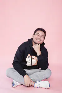 Pael smiling in a black hoodie while sitting cross-legged against a pink backdrop
